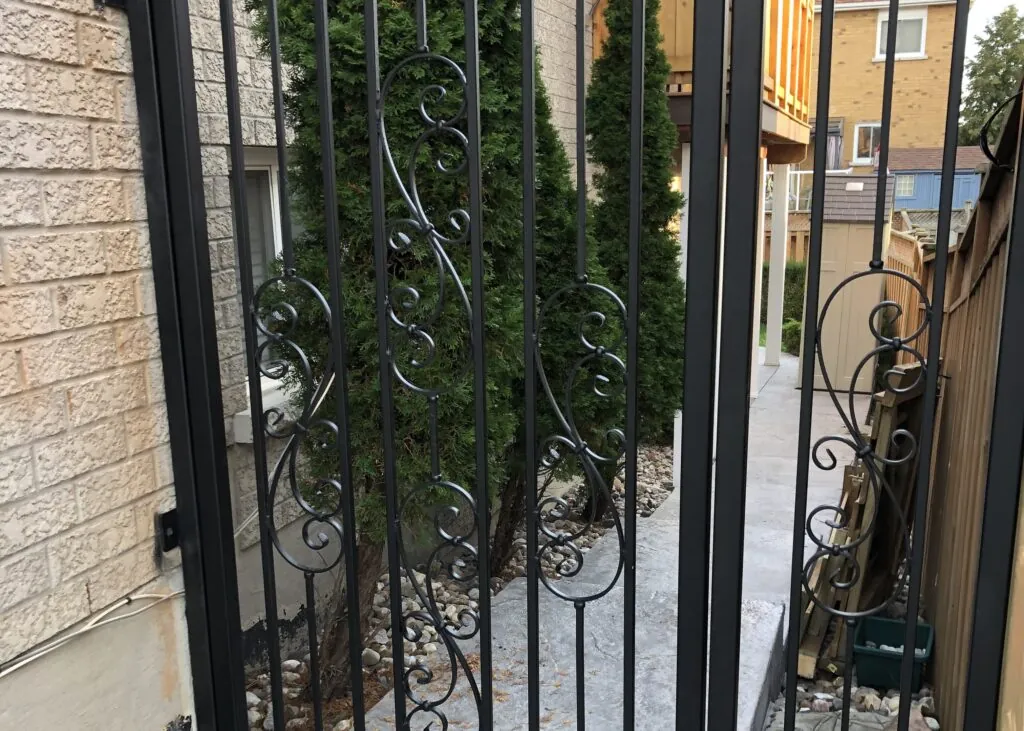 Black wrought iron gate with scrollwork details leading to a side walkway lined with cedar trees next to a brick house.