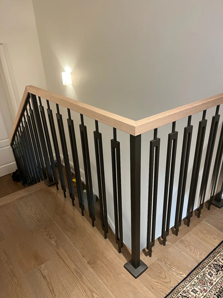 Black metal railing with decorative vertical balusters and a light wood handrail around an interior stair opening
